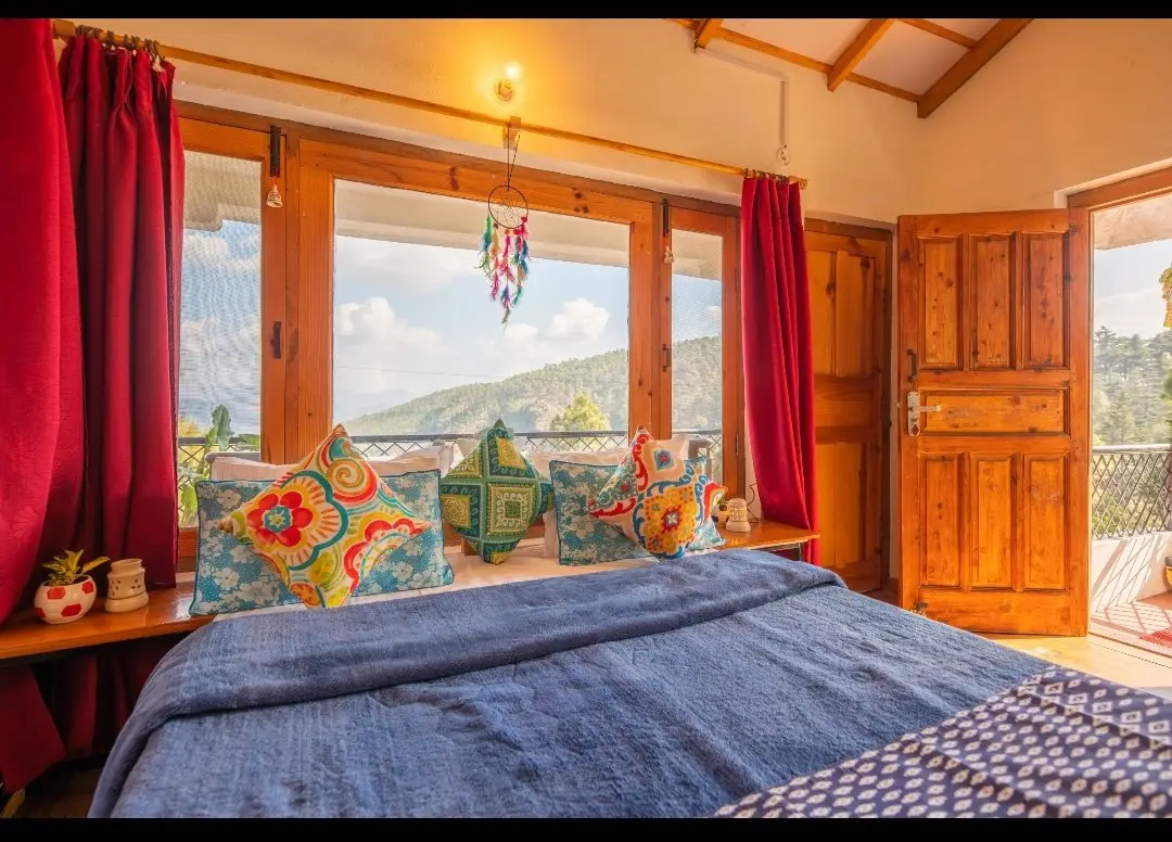 A bed with colourful cushions under a wide window, door open to the balcony