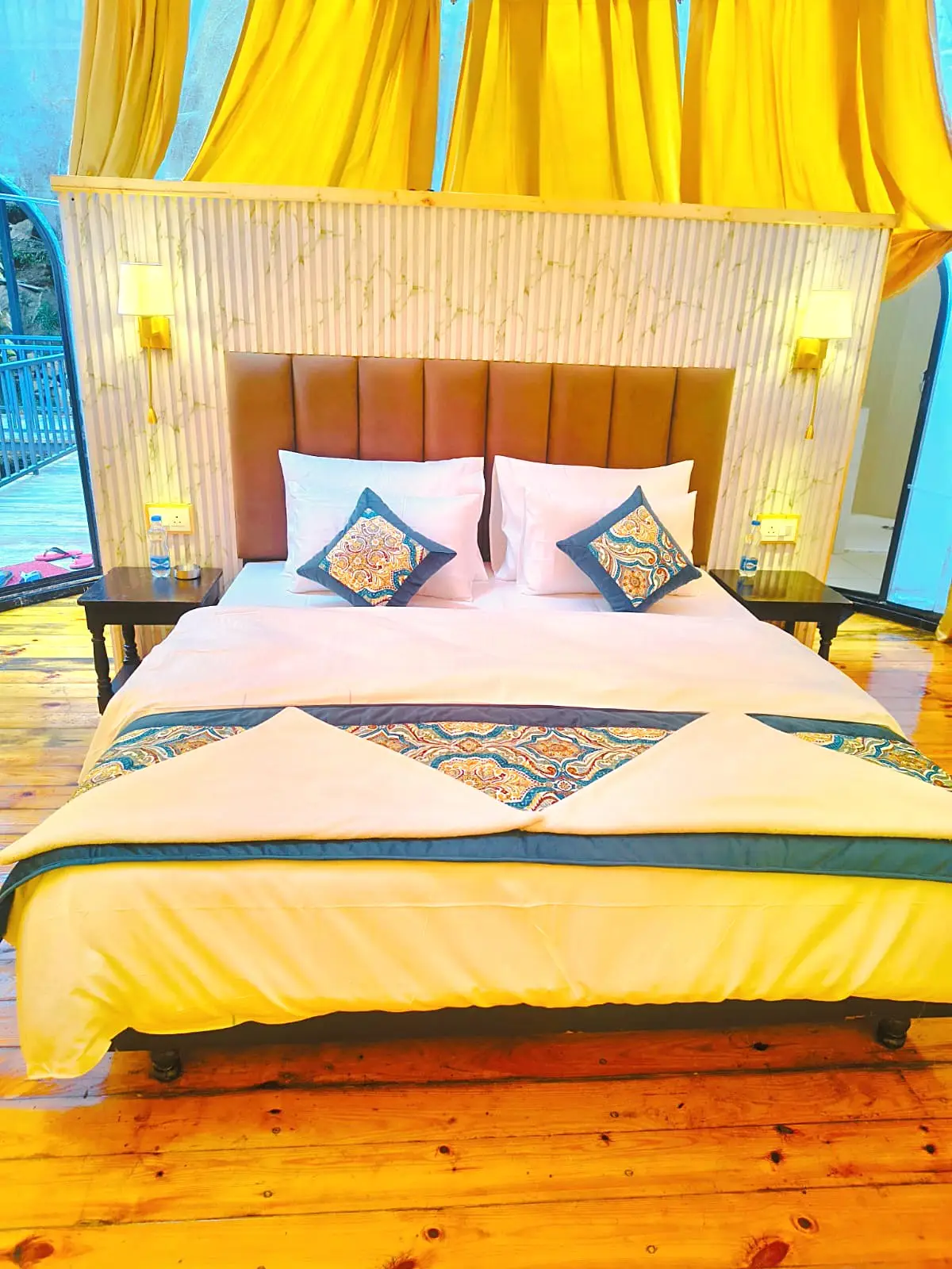 A double bed with a padded headboard, bedside lamps and yellow curtains