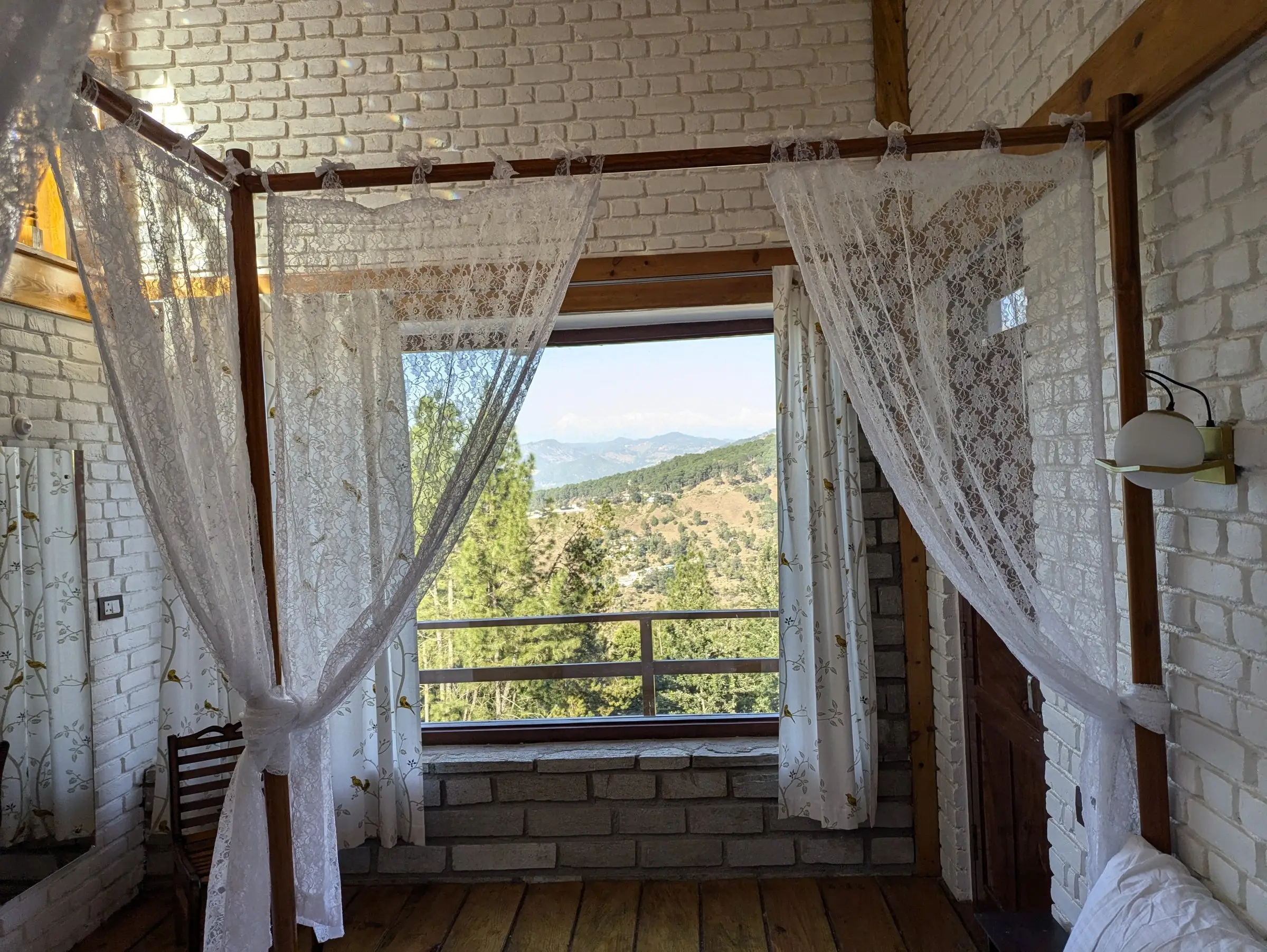 A window framed by lace curtains, looking over pines to distant hills