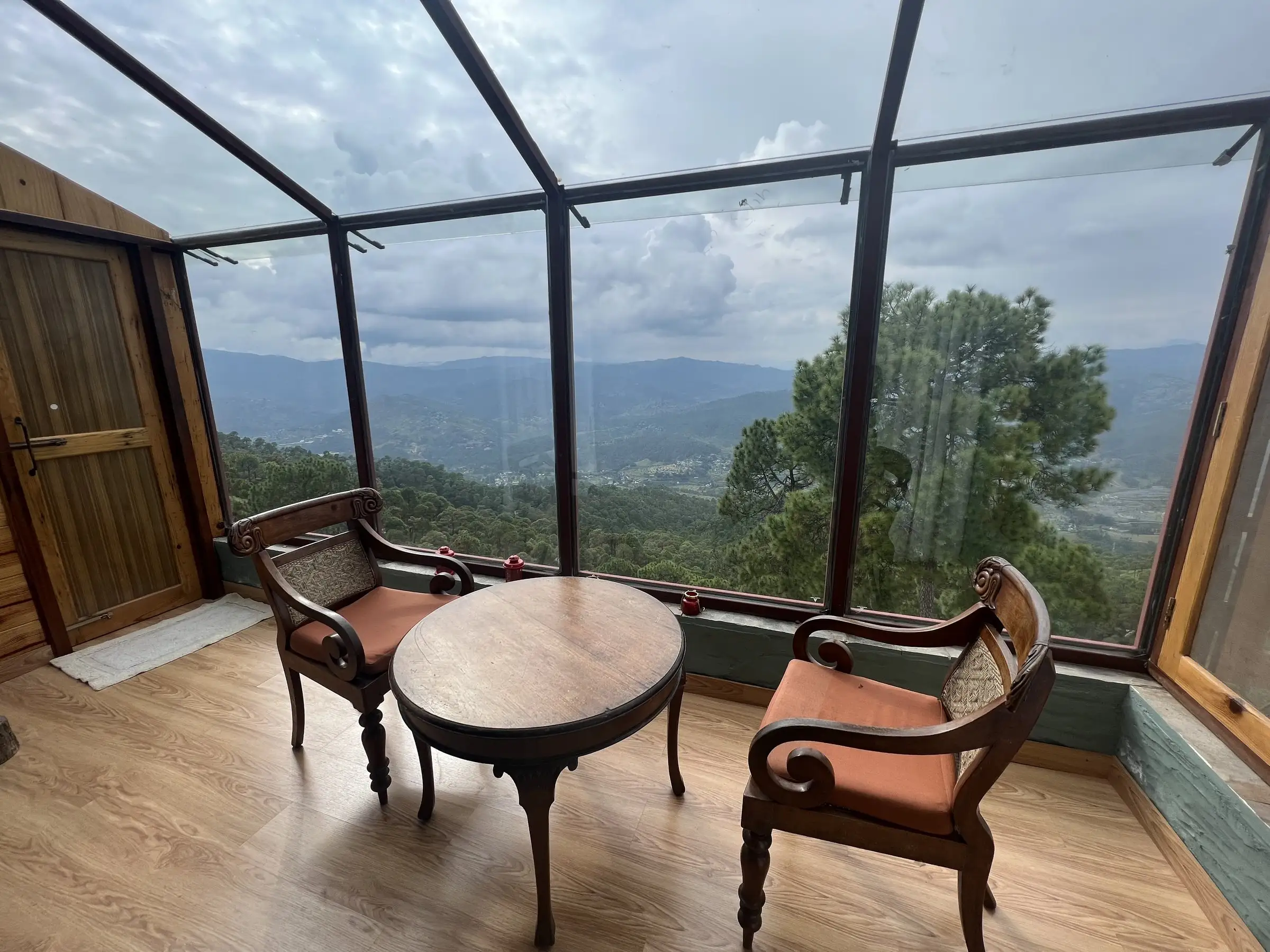 A glass-walled corner with a round table and two chairs over a forested valley