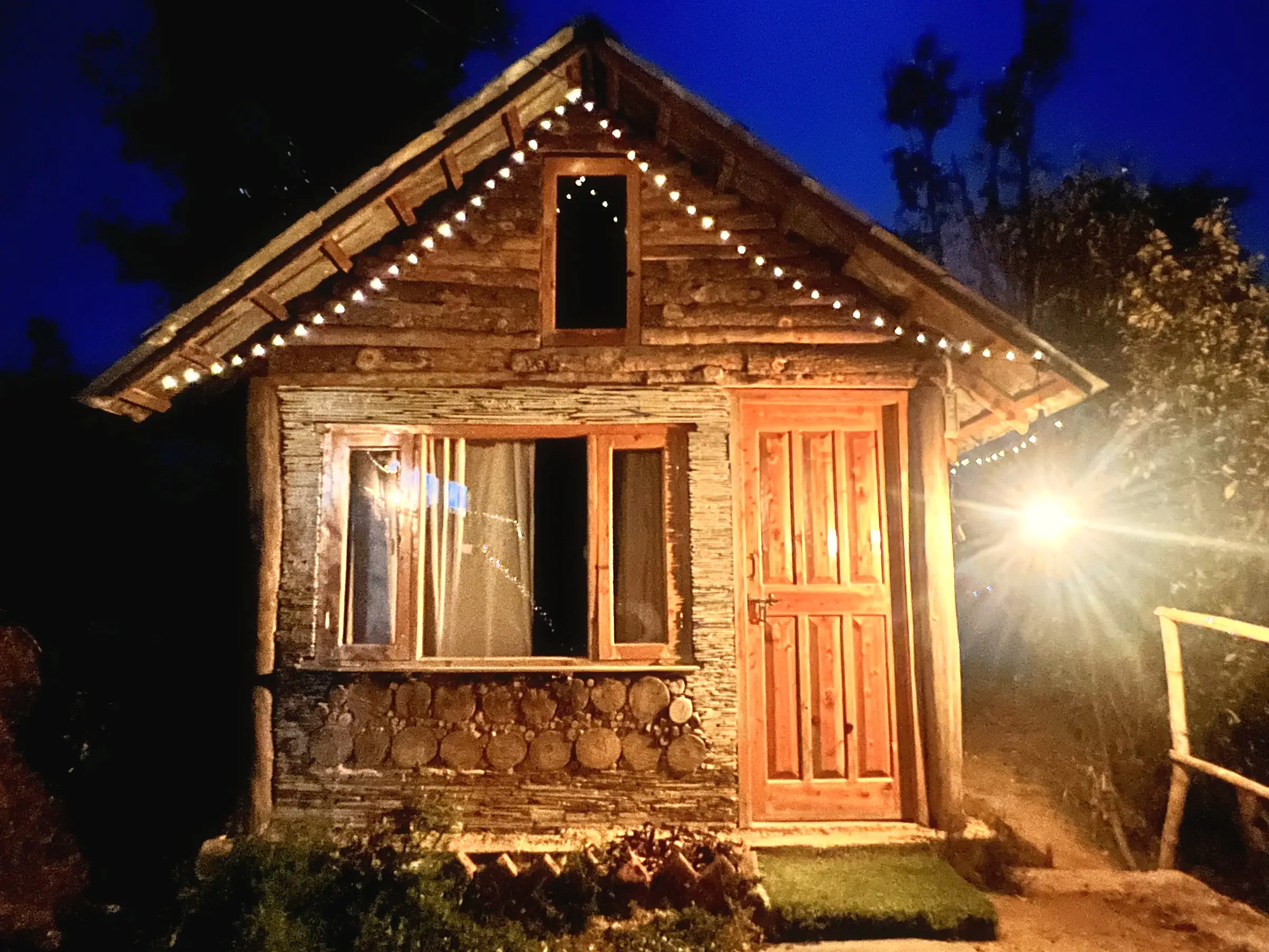 A small log-and-stone cabin at night, with string lights along the roof