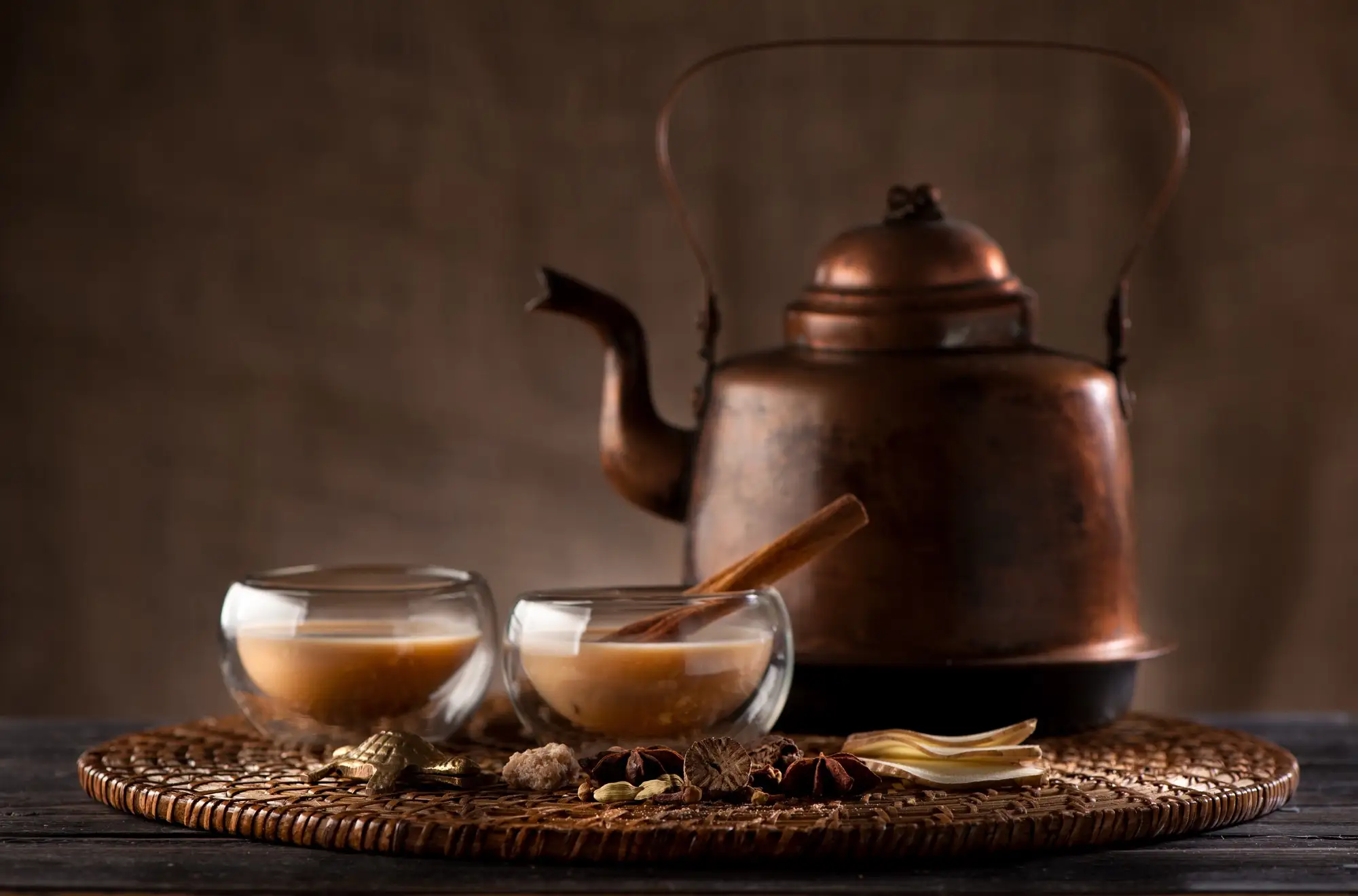 Glasses of spiced chai with a kettle and whole spices on a dark table