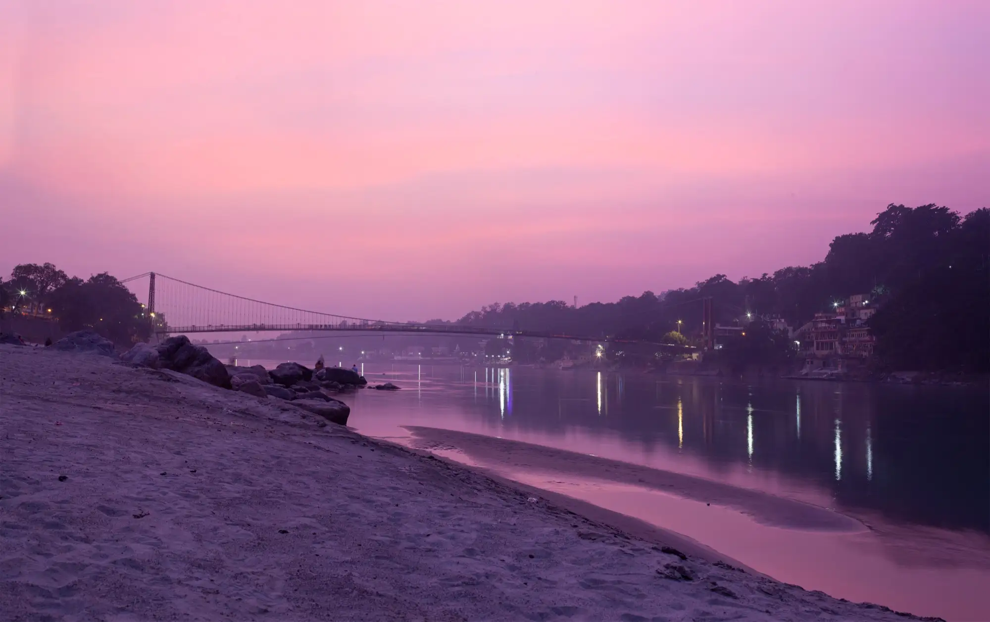 The Ganga at Rishikesh under a pink evening sky, with lights along the bank