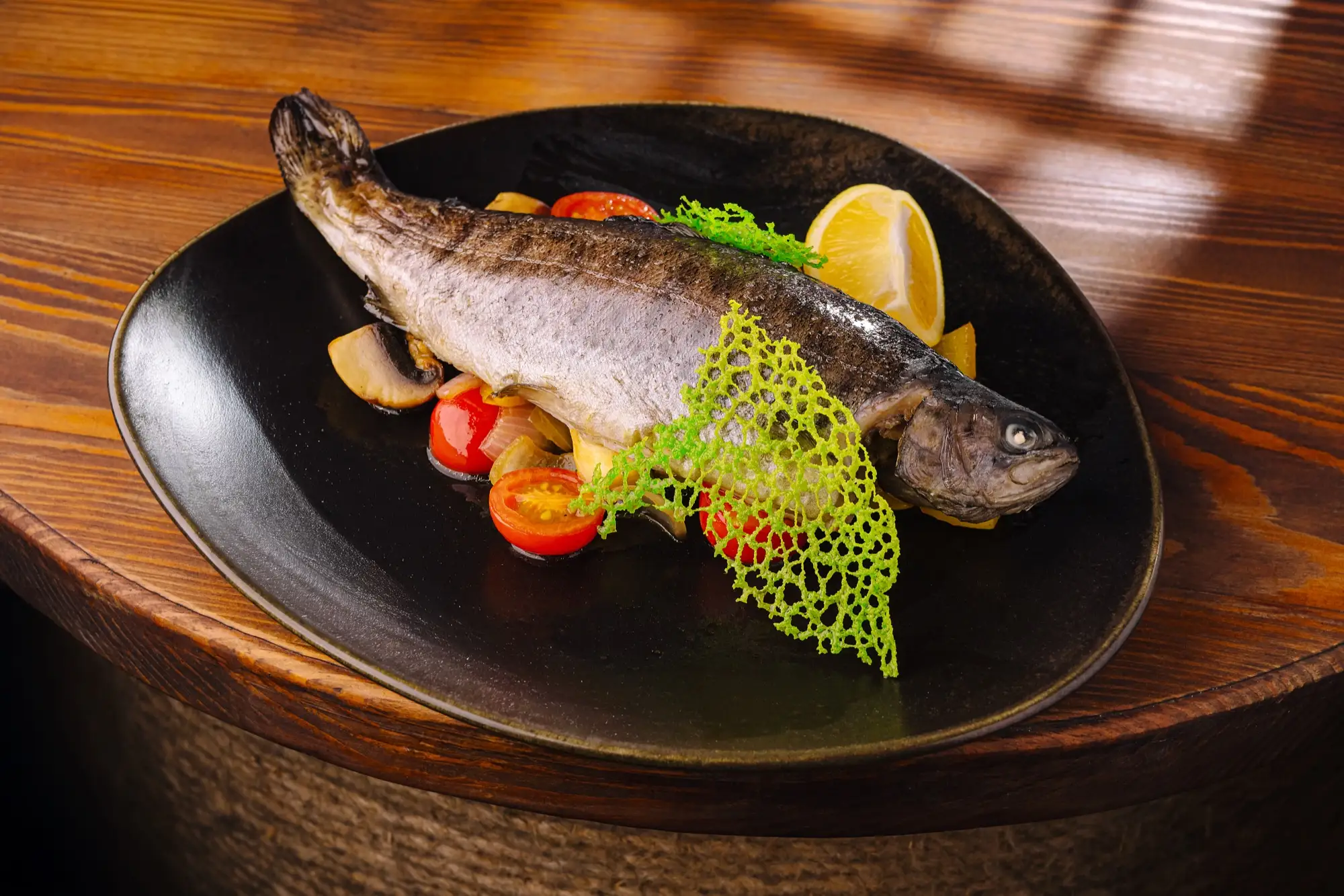 A whole grilled trout on a dark plate with roasted vegetables and lemon