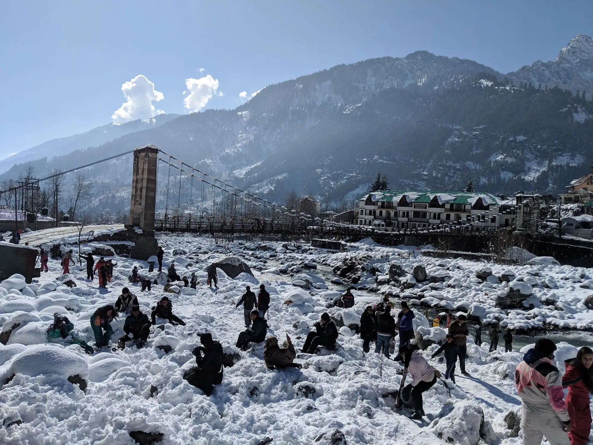 Visitors playing in fresh snow on the Beas riverbank in Manali, with a suspension bridge and hillside buildings behind